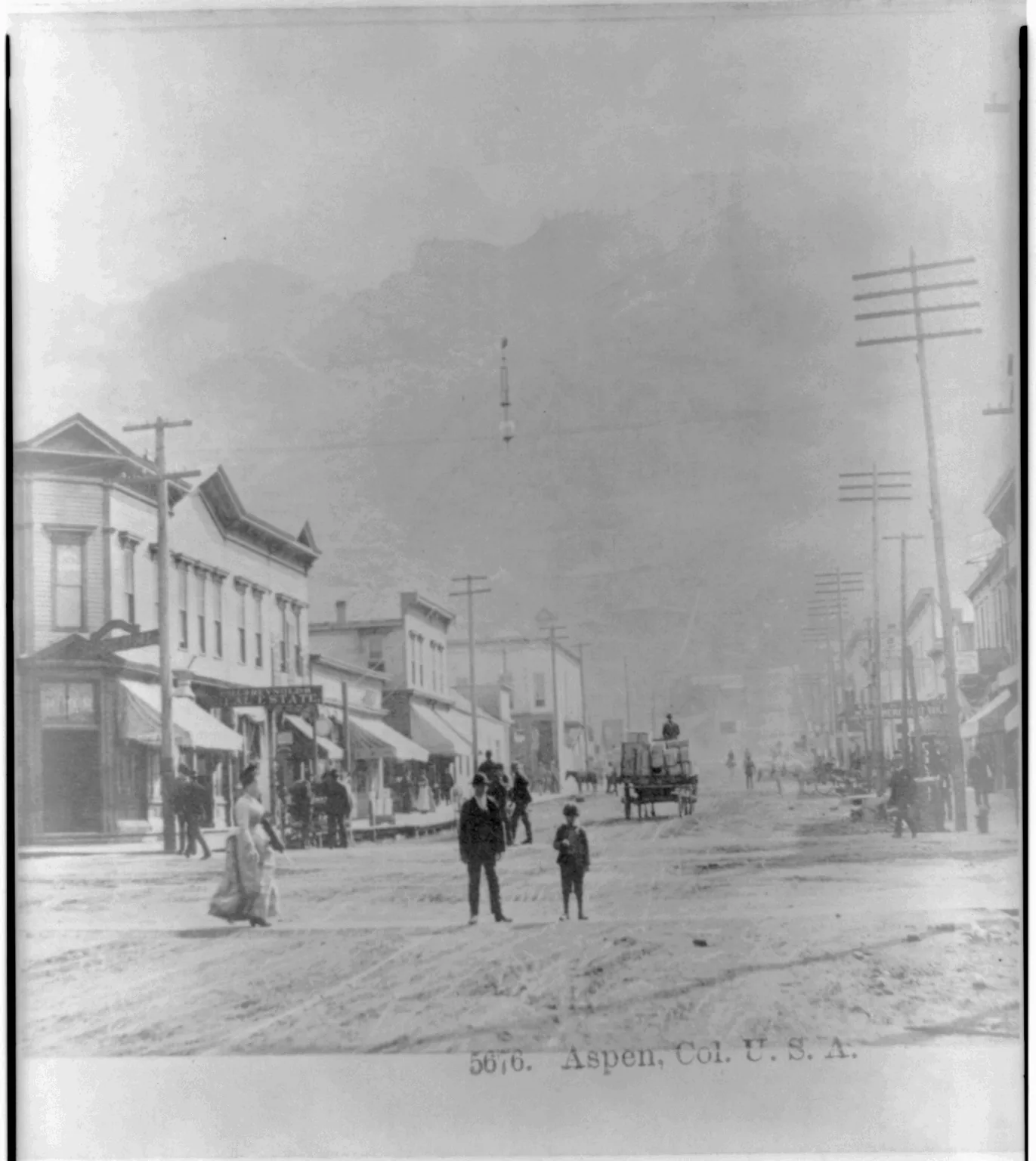 A wide dirt main street in Aspen, Colorado, photographed in 1890. Two-storey false-front brick and frame buildings with awnings line the left side, telegraph poles run down both sides toward a horse-drawn freight wagon, a woman in a long dress walks a child along the near edge, and a man and a boy stand in the middle of the road. A mountain face rises through the haze at the end of the street.
