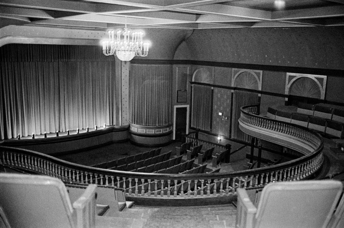 The Wheeler auditorium from the balcony, with the curved rail in front, rows of orchestra seats below, and the draped proscenium beneath a cut-glass chandelier.