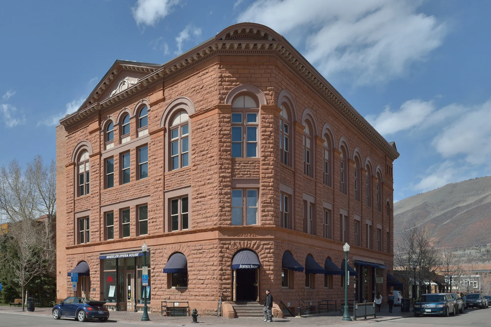 The Wheeler Opera House on its street corner: three storeys of rusticated peachblow sandstone, round-arched windows on the top floor, a dentil cornice and pedimented gable, navy awnings over the shopfronts at street level, with Aspen Mountain rising behind it.