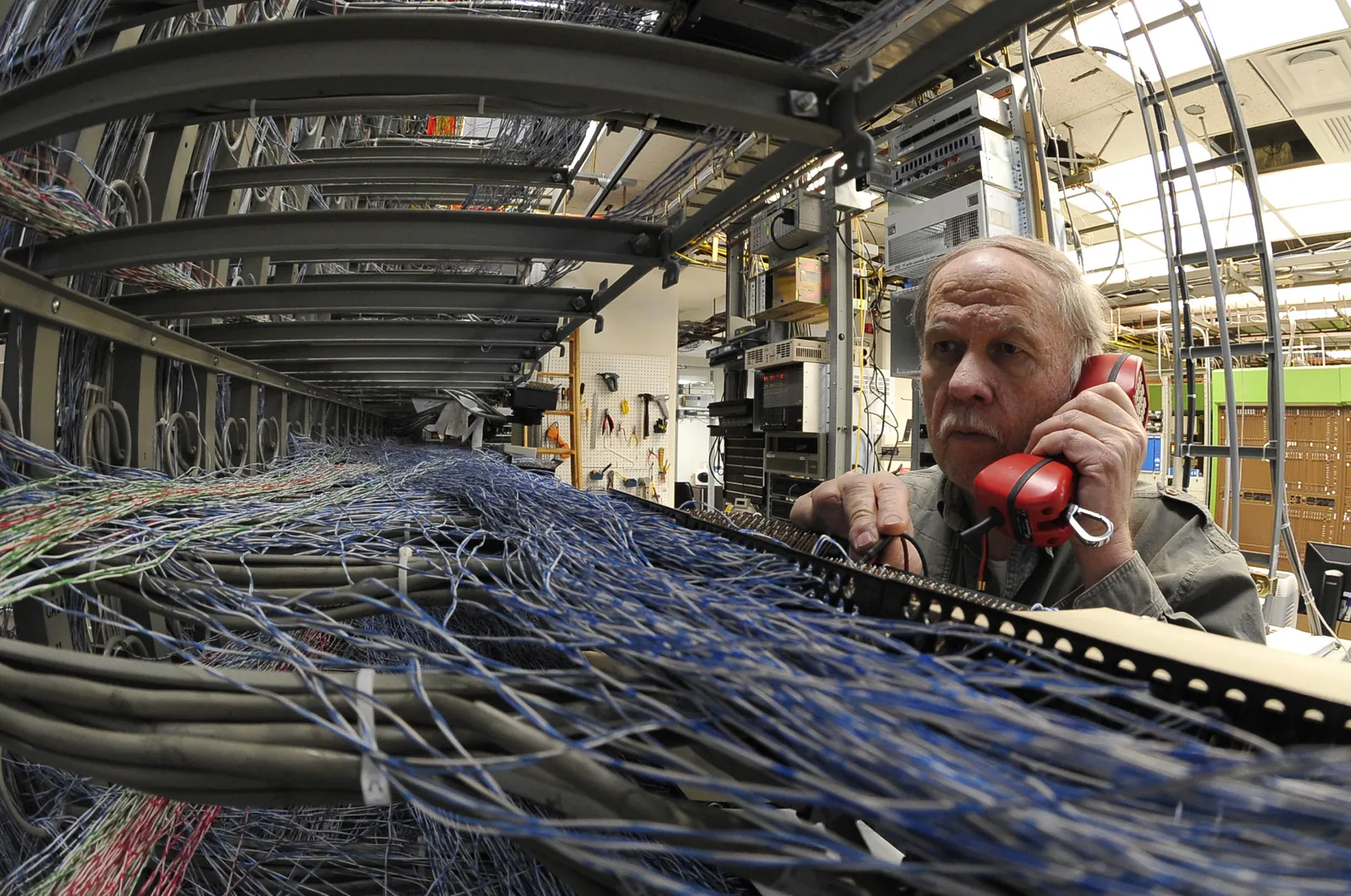 A technician holds a red telephone handset to his ear while checking for dial tone at a telephone switch, with blue cross-connect wiring spread across the frame in front of him.