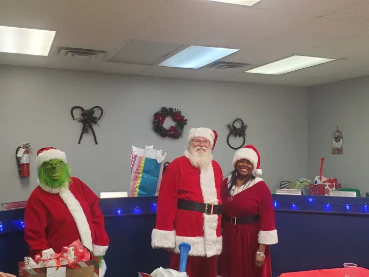 The Grinch, Santa Claus and Mrs. Claus stand together at a counter, holiday wreaths hung on the wall behind them and gift bags set out beside them.