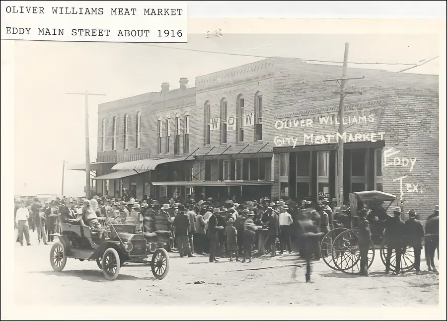 A crowd of about a hundred people in hats fills the dirt street outside the Oliver Williams City Meat Market, with a Model T at the left and a horse-drawn wagon at the right.
