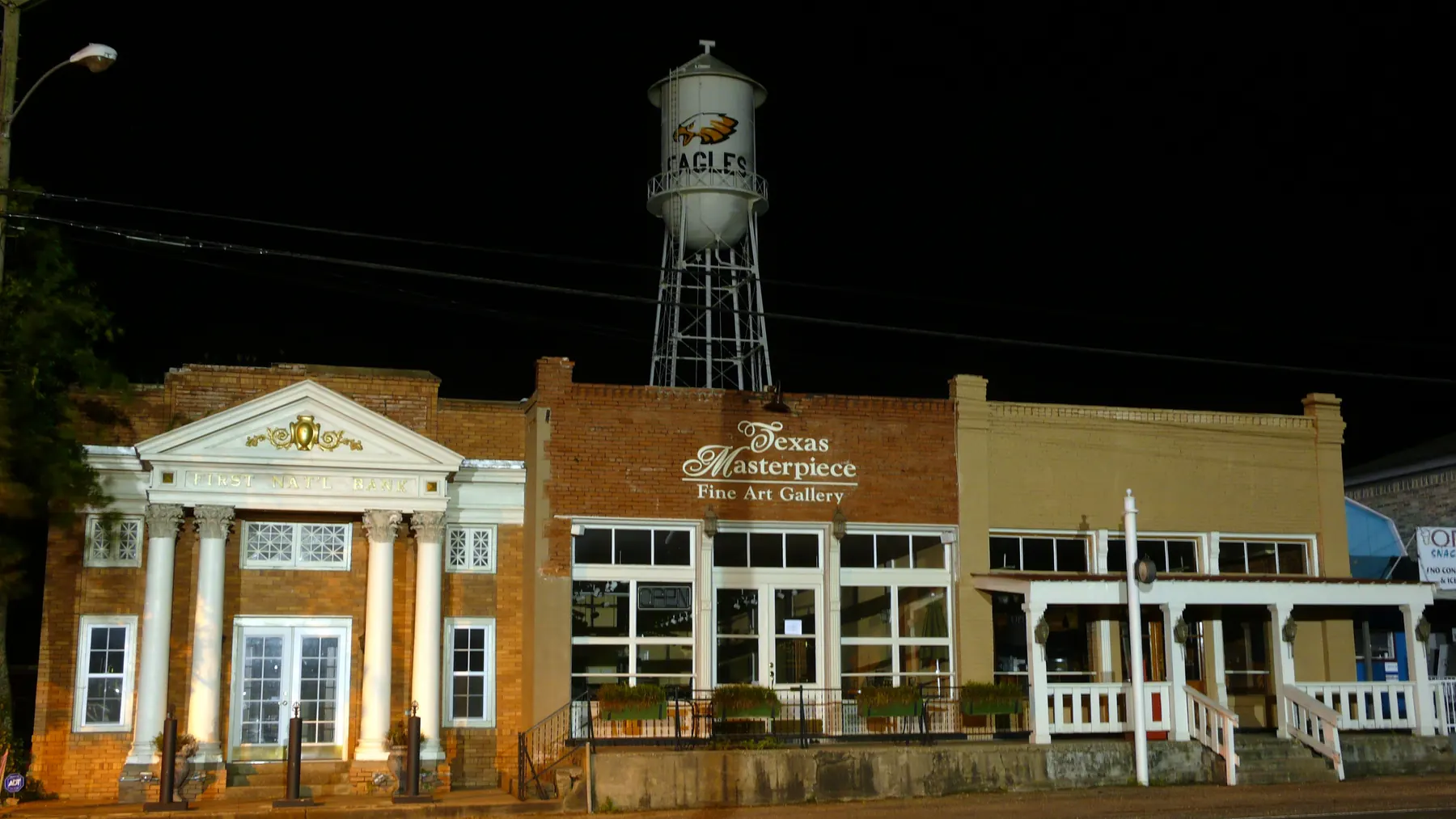 The Eagles water tower lit above the brick storefronts of Eddy Main Street at night, with a columned bank building on the left and the Texas Masterpiece gallery in the middle of the row.