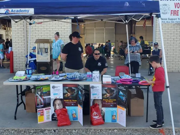 Volunteers hand out information and prizes from tables under a canopy at Community Night Out while families walk through.