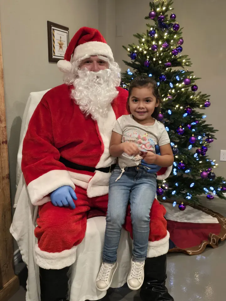 Santa Claus and a small child sit together beside a Christmas tree strung with blue and purple lights.