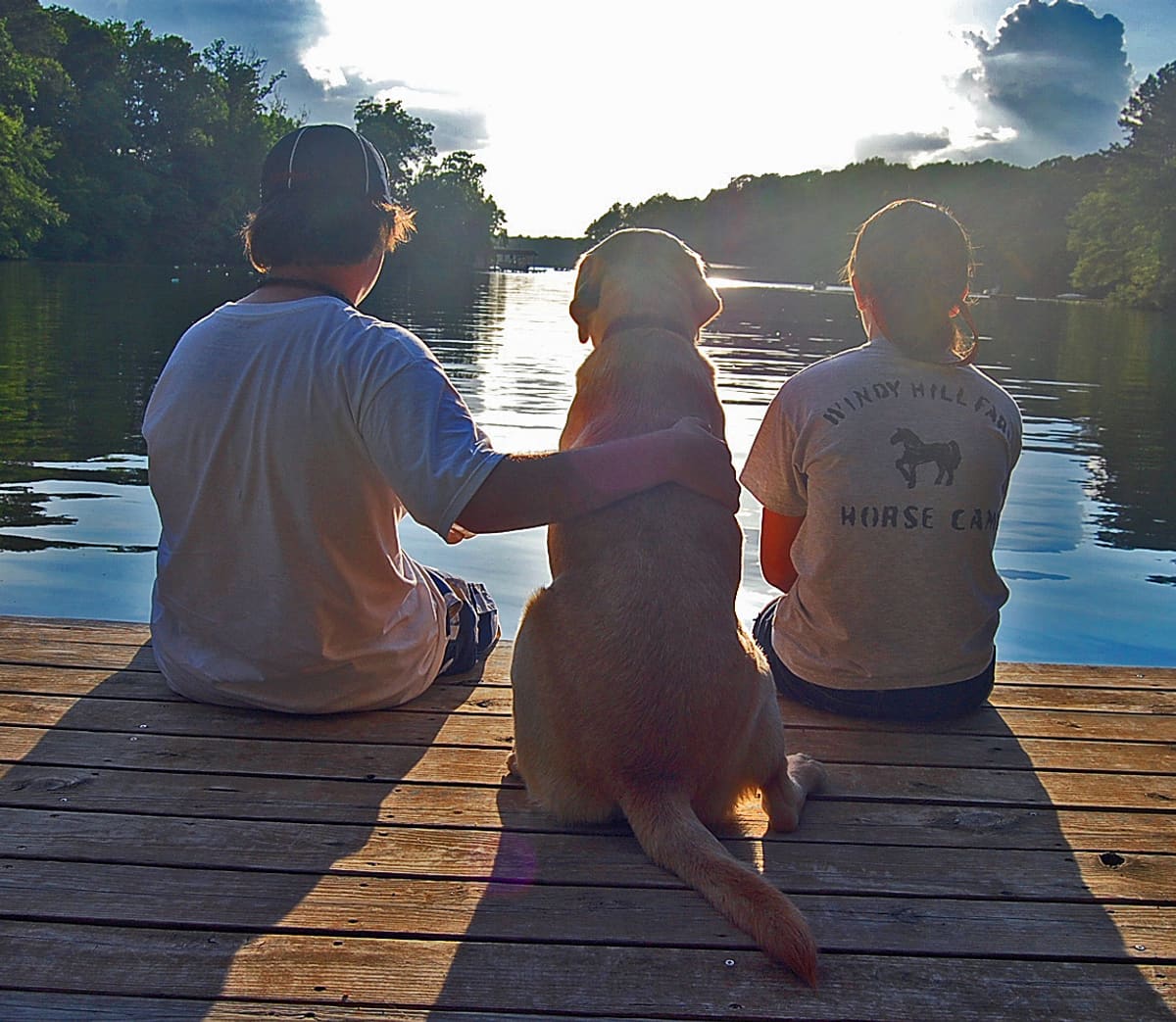 Two people and a yellow dog sitting together at the end of a wooden dock on Lake Gaston, looking out at the water with the sun low over the far treeline