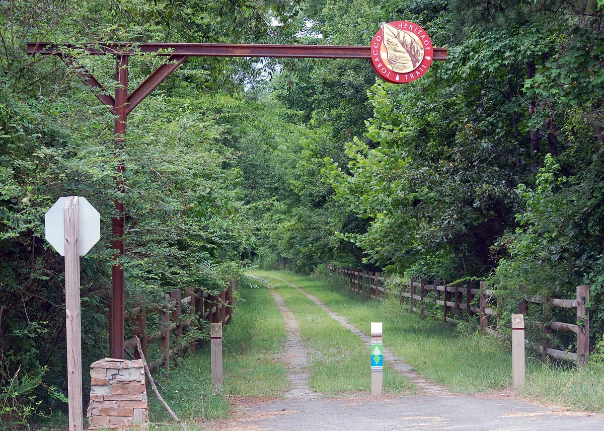 The rusted iron gateway over the Tobacco Heritage Trail, hung with a round red trail sign, where the old rail bed runs on between split-rail fences