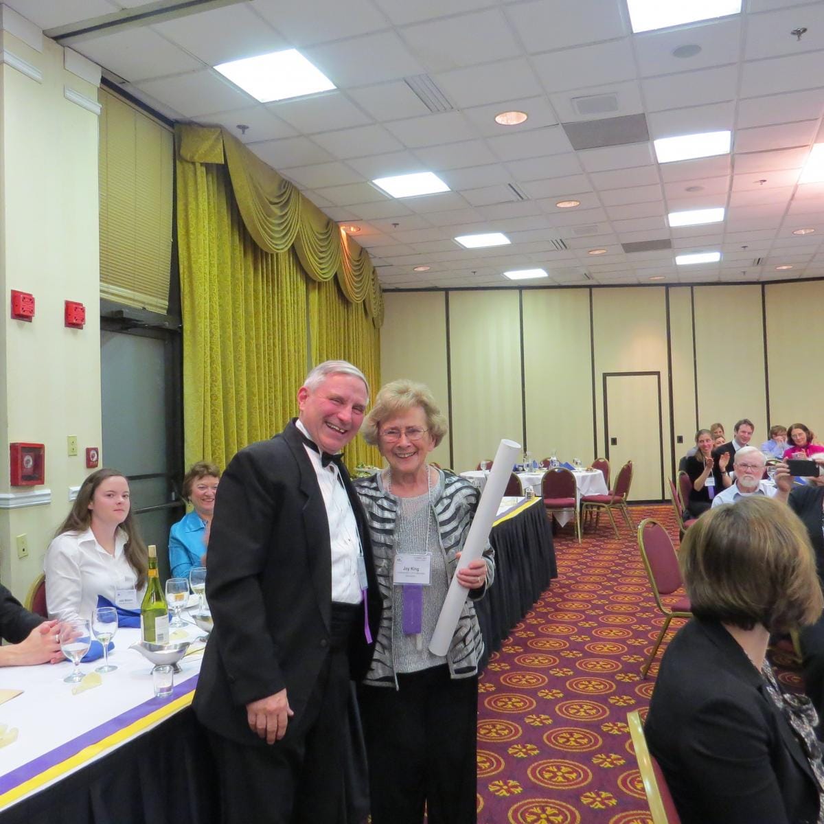 Joy King and James May with her ovatio scroll at the CAMWS banquet, with diners at tables behind them