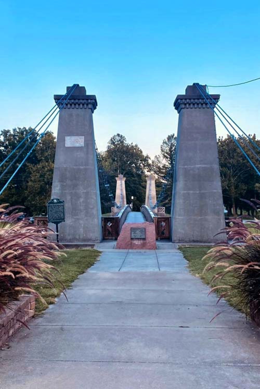The two stone towers of the General Dean Suspension Bridge framing the wooden walkway across the Kaskaskia River, with a historical marker at the entrance.