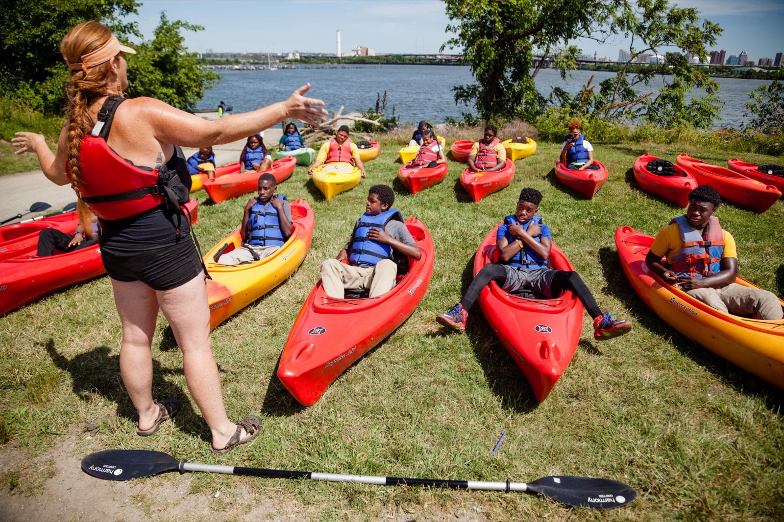 Students in life jackets sit in kayaks on a grassy bank while an instructor teaches beside the water