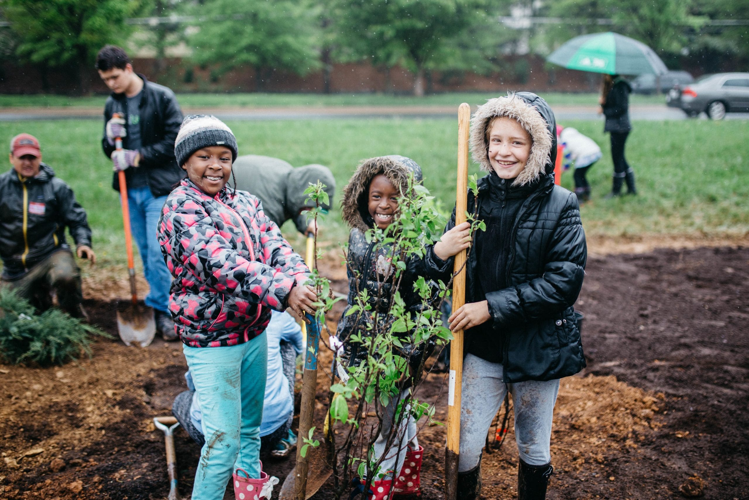 Three children smile while planting saplings in turned soil on a rainy day