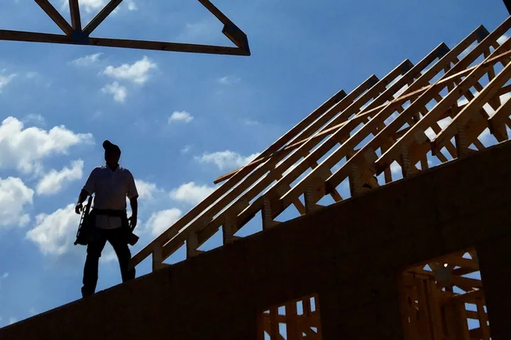 A construction worker standing on the wall plate beside new roof framing, with a suspended truss entering the frame.