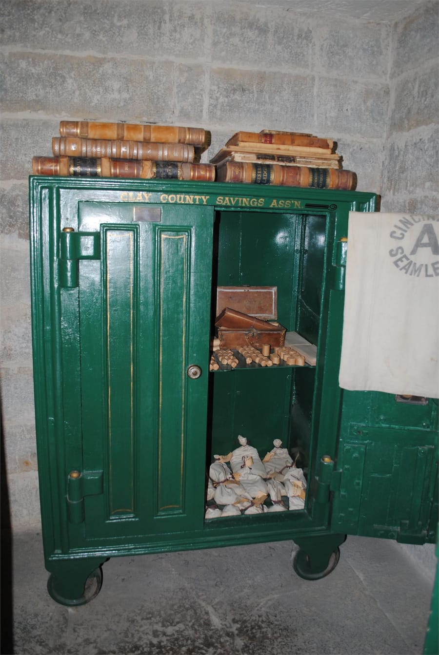 A green iron safe with its doors open, stacked with ledgers and coin bags.