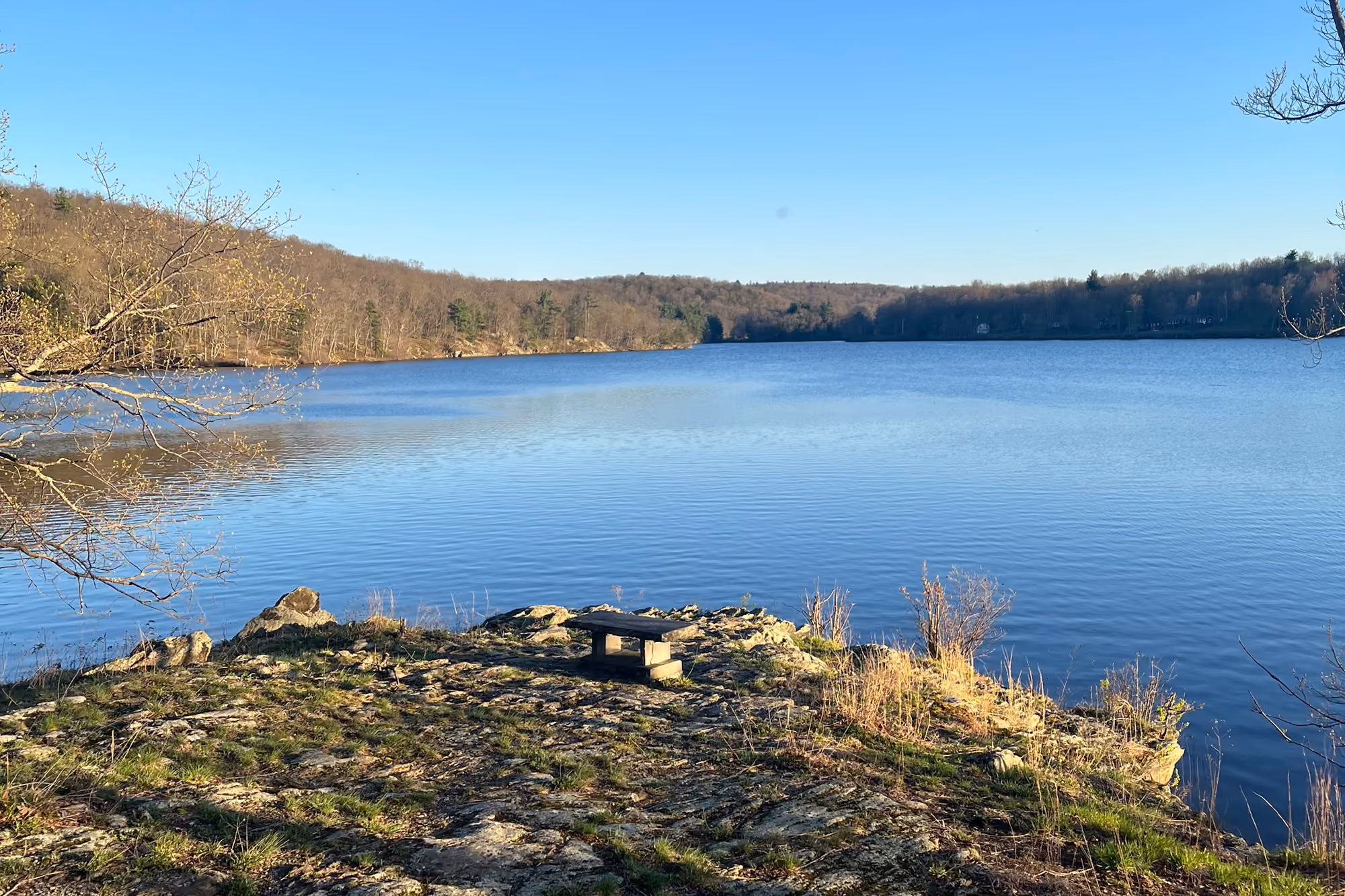 Lake Taghkanic under a clear blue sky, a bench on a rocky ledge in the foreground and wooded hills along the far shore