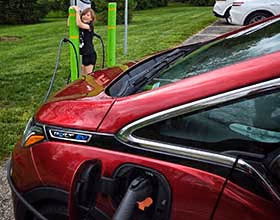 A red electric vehicle hooked up to an EV charging station in a public park. A young girl watches in the background.