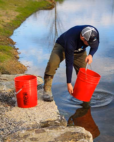 A man with two red buckets with one foot in a pond and the other foot on a rock at the pond's edge.