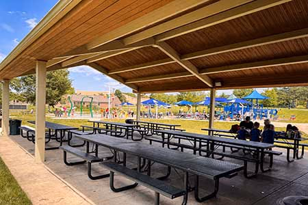 The inside of a picnic shelter. One table is in use by a family. Splash pad and playground can be seen in the background.