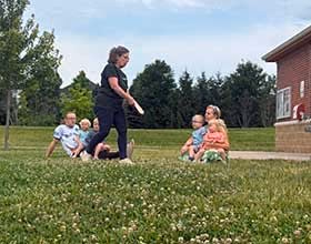 A person is standing in the grass. Several people are seated around her.
