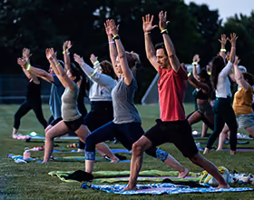 Image of multiple people participating in an evening outdoor yoga class in a park grassy field. They stand on towels and yoga mats. Everyone is standing in a warrior pose, lunging with one knee and arms stretched up above their heads. Everyone wears glow bracelets.