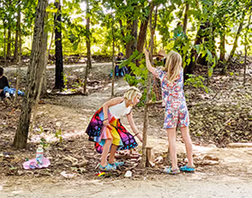 Image of two children building a fairy house in the woods. One wears a colorful, butterfly-inspired skirt. The other wears a floral print outfit. One child squats to add to the fairy house, the other is tying string to a branch of a tree.