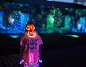 A child in a bright pink shirt wearing glow-in-the-dark glasses and a necklace. The background is mostly dark with some blue and green light illuminating a brick wall.