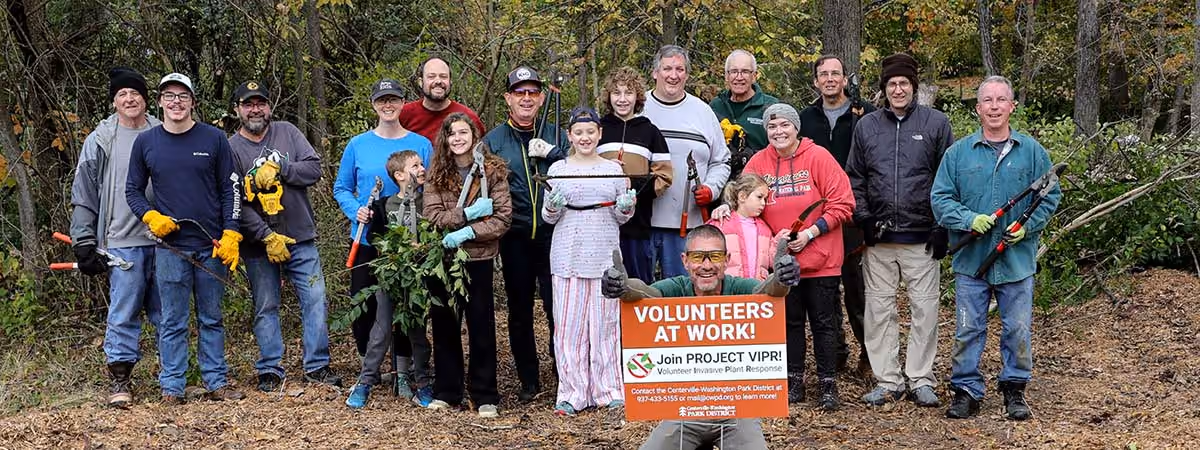 A group of people standing outside. One man gives two thumbs up while kneeling behind a “Volunteers at Work” sign