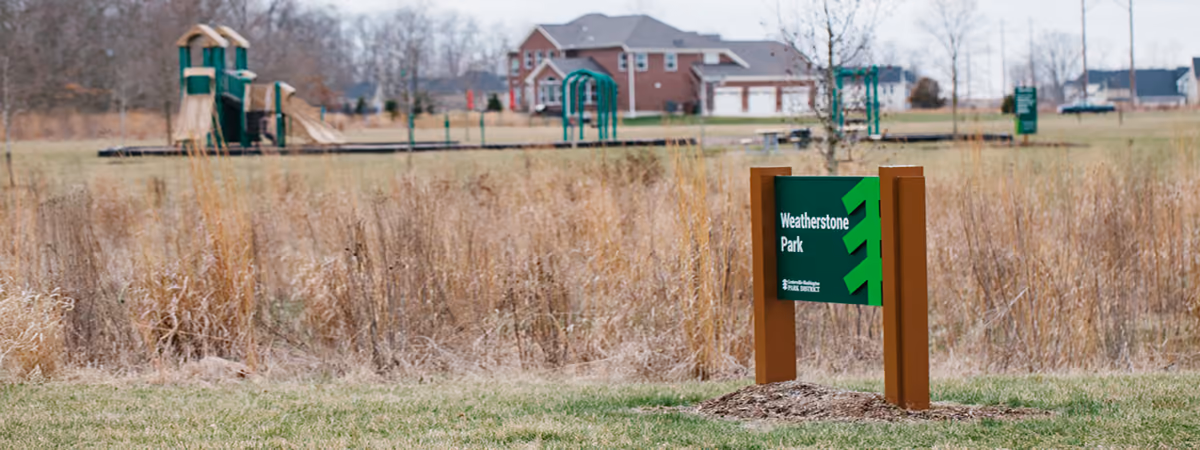 The Weatherstone Park sign in the prairie grasses, with the playground behind