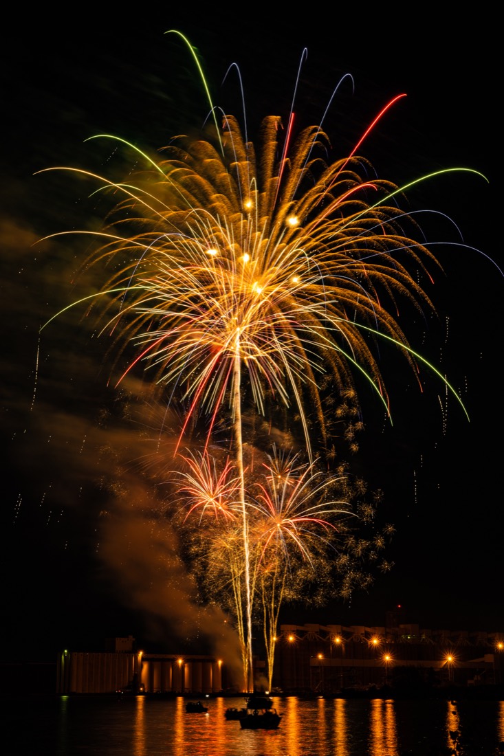Fireworks bursting over the Duluth harbor at night, with grain elevators silhouetted below