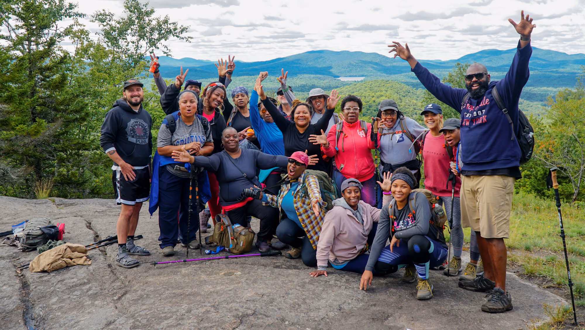 A hiking group celebrating with arms raised on a bare Adirondack summit, mountains behind