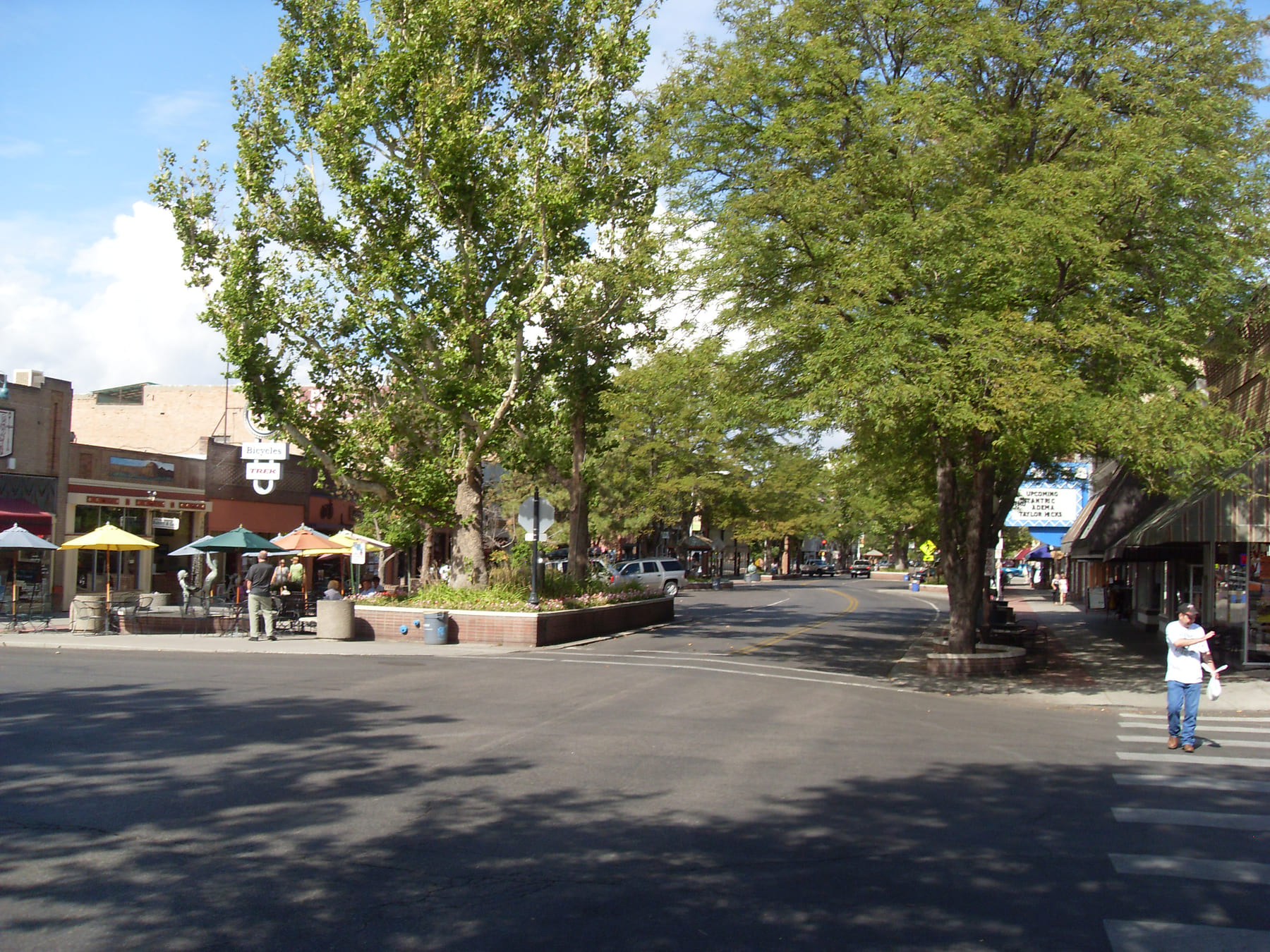Main Street looking down its length: the roadway curves around planted brick medians full of mature shade trees, with shopfronts and awnings on both sides, café tables and umbrellas on the left pavement, and a figurative sculpture standing beside them