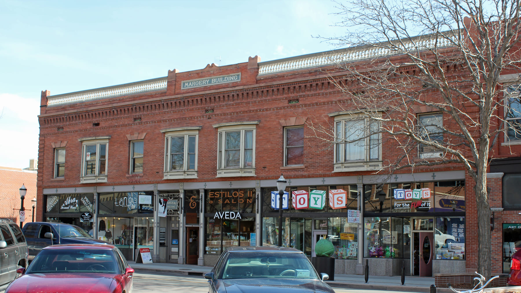 A two-storey red-brick commercial block on Main Street with its name carved into the parapet, four shopfronts along the pavement beneath it, a salon and a toy shop among them, and cars parked at the kerb