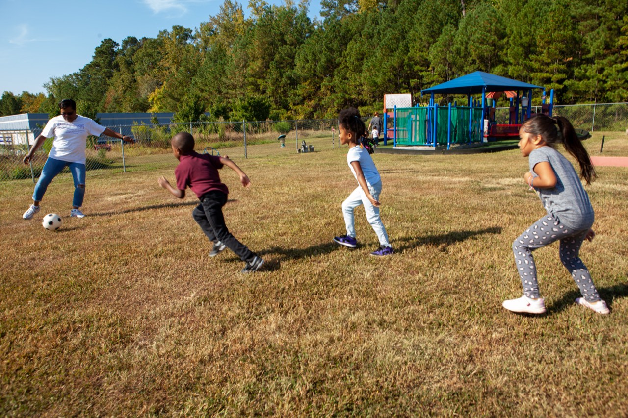 Durham PreK children playing soccer outdoors with a teacher beside a school playground