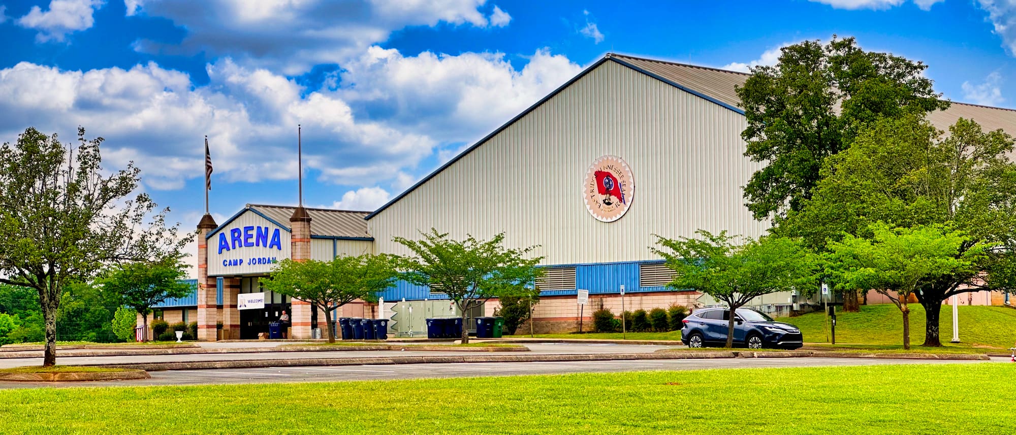 Camp Jordan Arena on a clear summer day: a long metal-clad hall with ARENA and CAMP JORDAN lettered above its brick entrance, a painted roundel high on the gable, young trees along the front, and mown grass in the foreground.