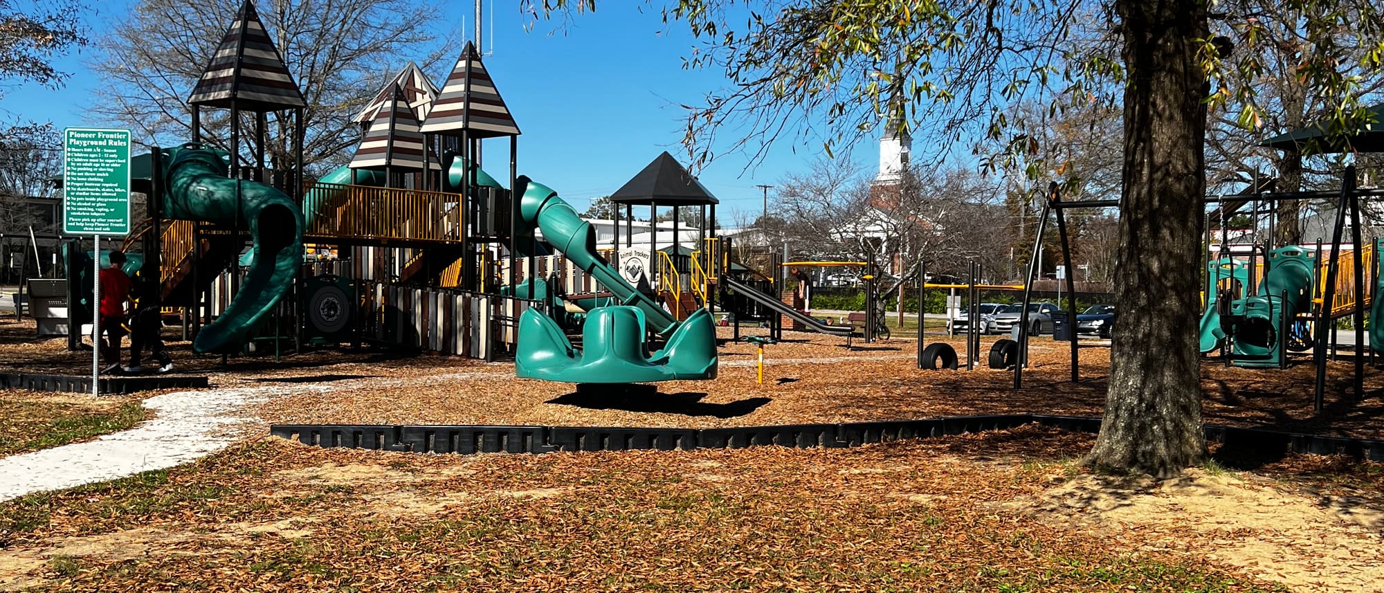 The Pioneer Frontier playground: timber towers with striped peaked roofs, green tube slides, swings and climbing frames over wood-chip ground, with a white church steeple beyond the trees.