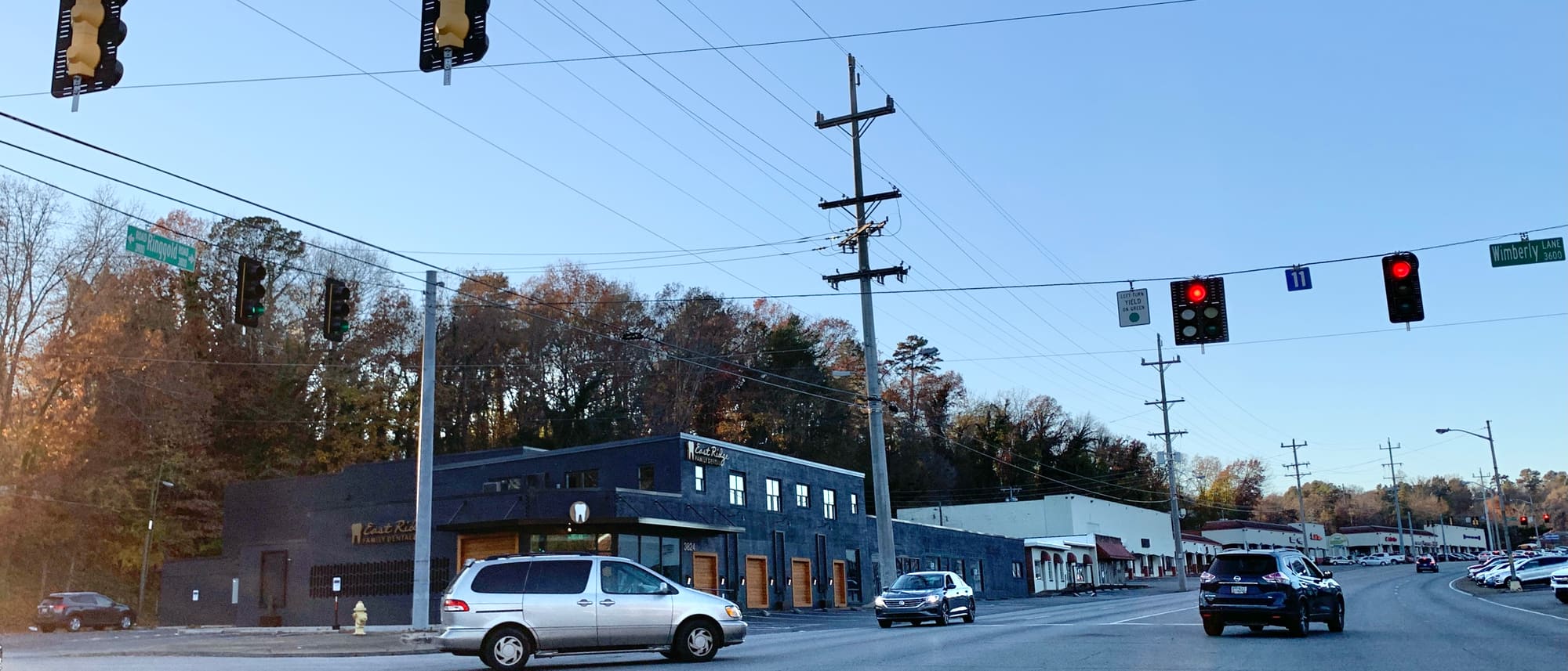 Ringgold Road at Wimberly Lane in late afternoon. A row of low brick and painted storefronts runs the length of the far side of the road, the wooded slope of the ridge rises directly behind them, and cars wait at a signal strung across the intersection.