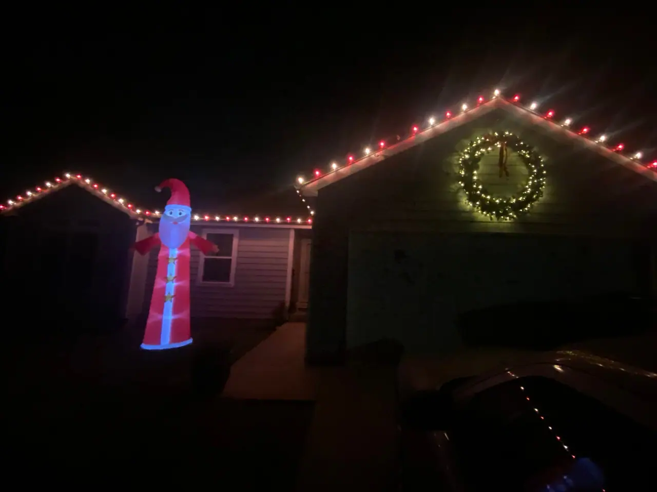 A house outlined in holiday lights at night, with a lit wreath and a lit figure in the yard.