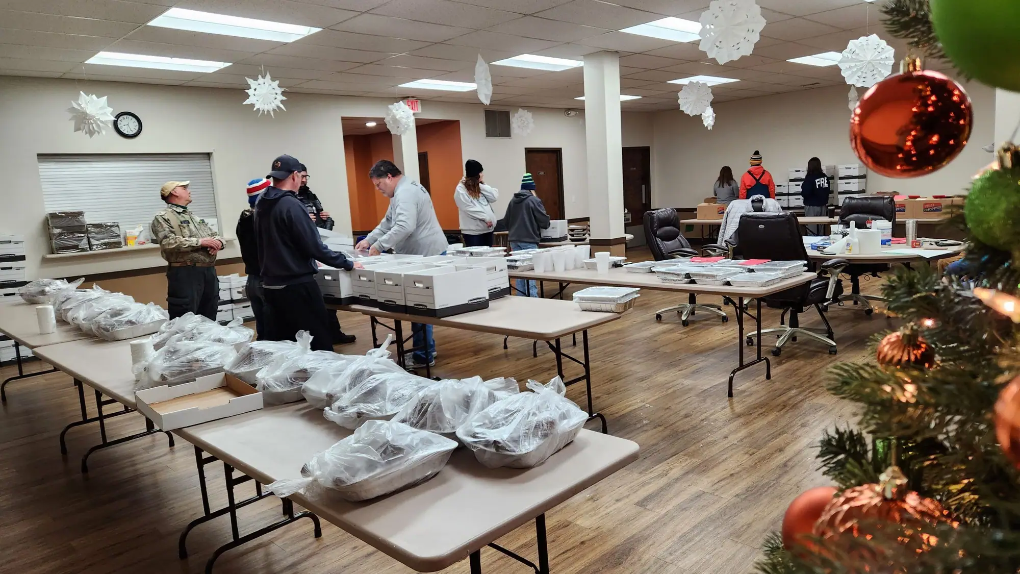 Volunteers set out bagged meals on long tables in a hall decorated with a Christmas tree and paper snowflakes.