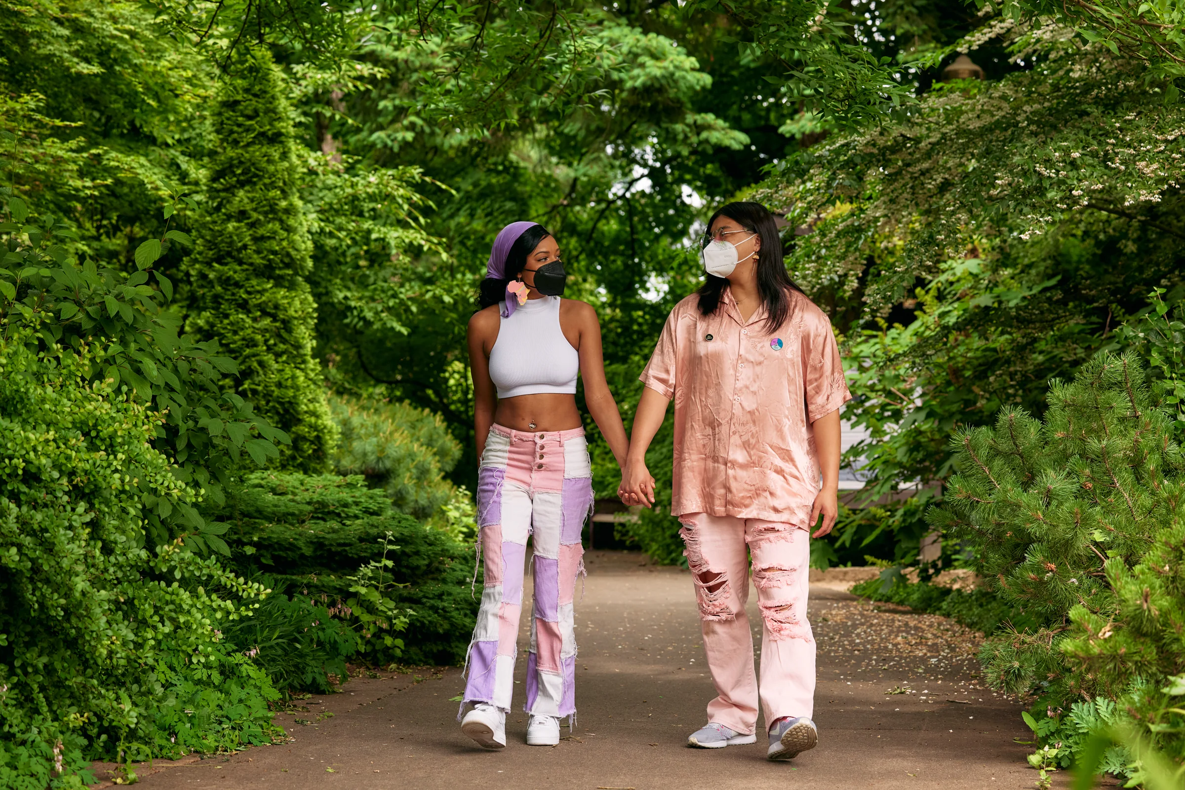 Two people hold hands while walking along a paved path through a leafy park.