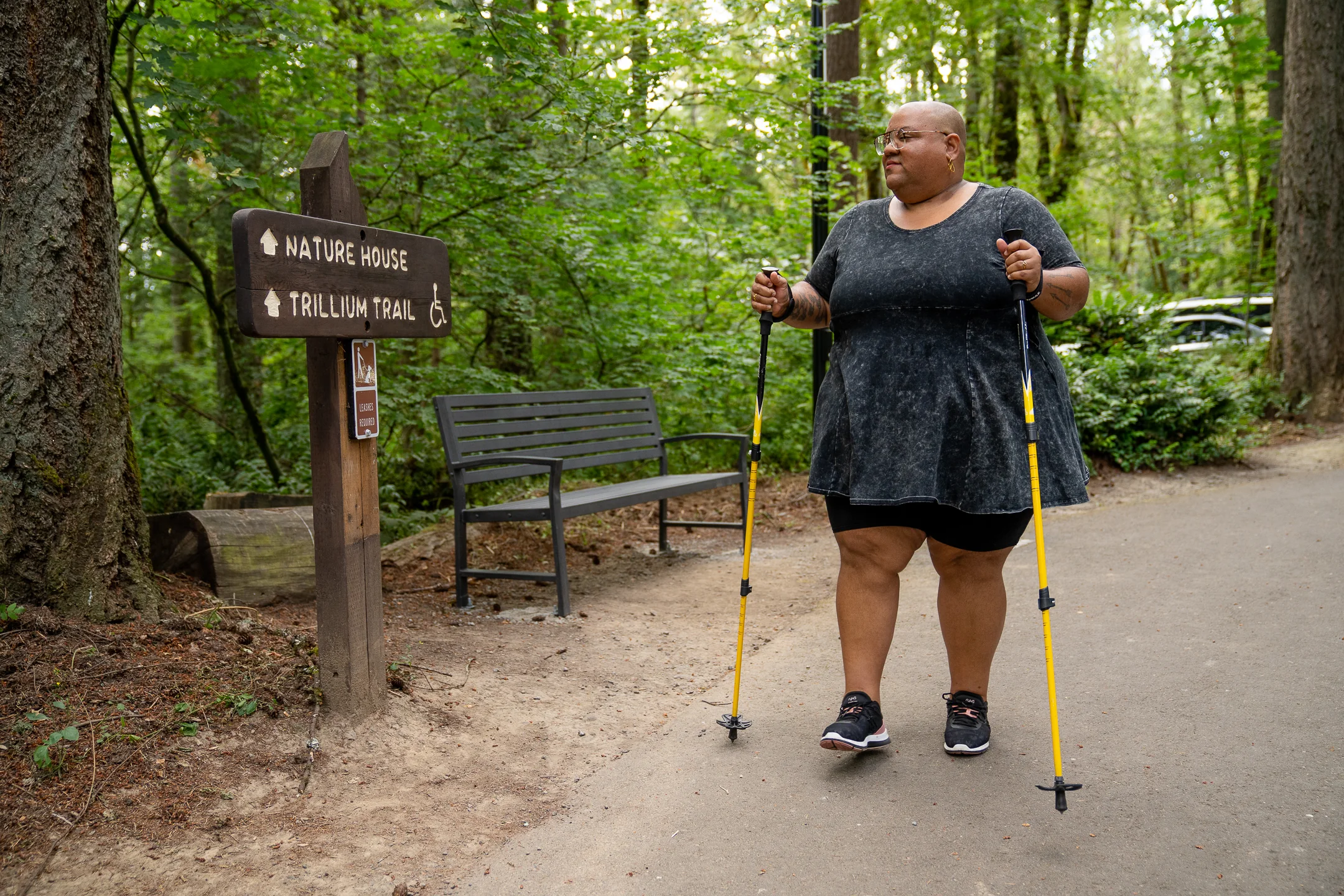 A hiker with trekking poles stands beside a trail sign marked with an accessibility symbol.