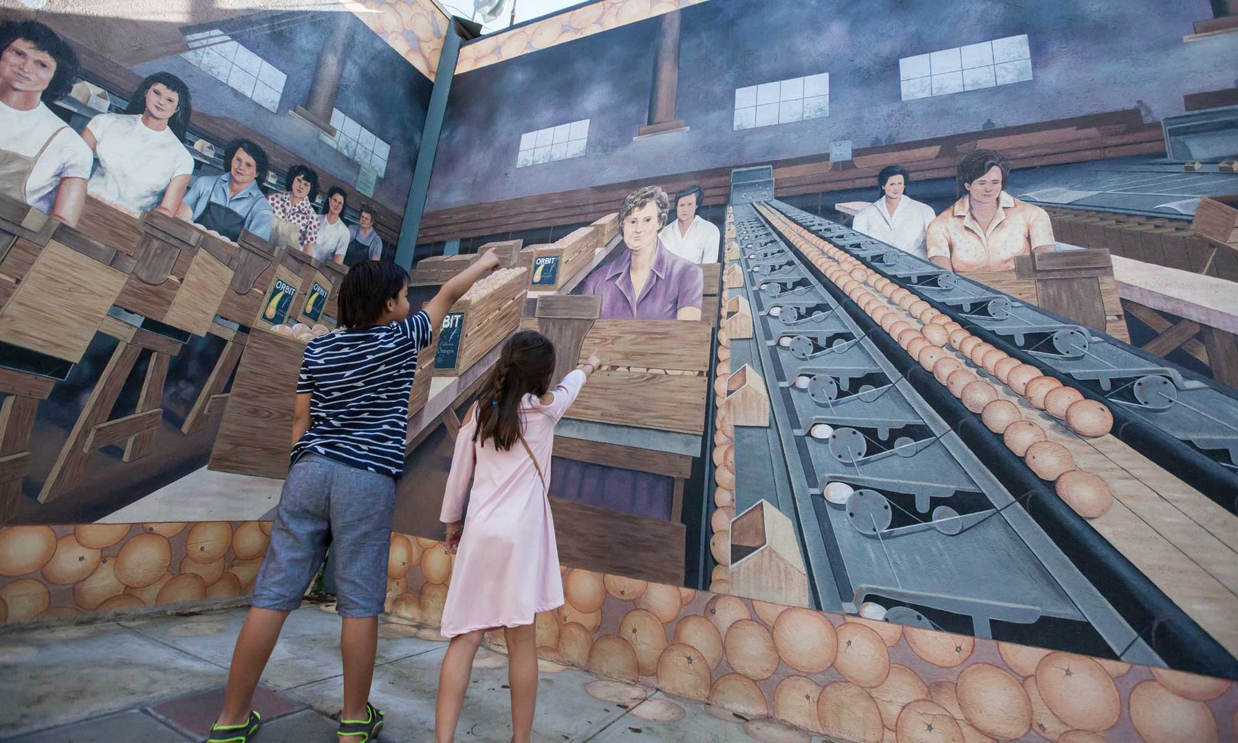 Two children reach up to touch a painted packing-house mural on a downtown Exeter wall, where women sort oranges along a conveyor.