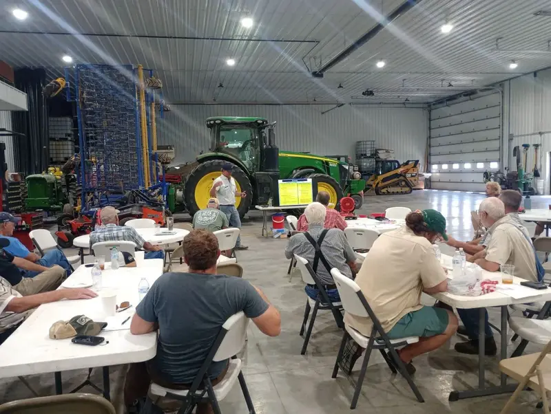 Farmers sit at folding tables in a large farm shop, with a green tractor and a planter parked behind them.