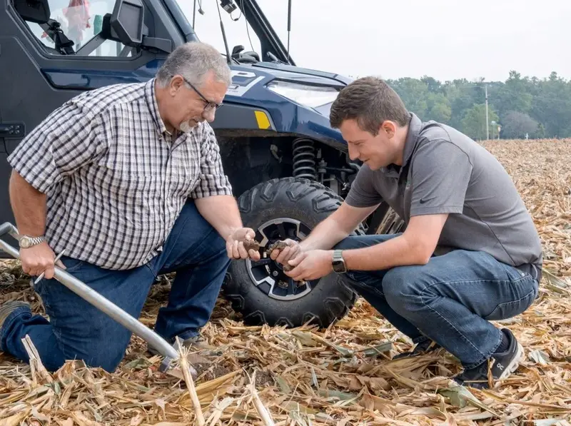 Rodney Rulon and his mentee Austin Conrad kneel in harvested cornstalks beside a utility vehicle, one holding a soil probe.