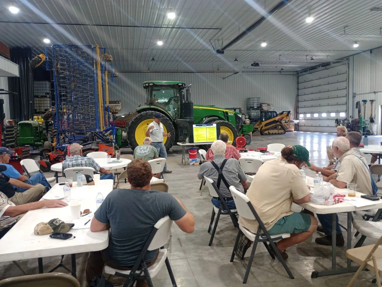 Farmers sit at folding tables in a large farm shop, with a green tractor and a planter parked behind them.