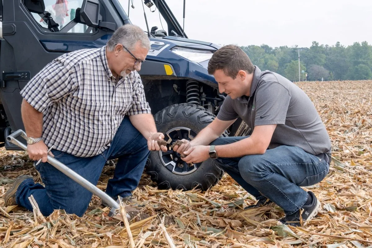 Rodney Rulon and his mentee Austin Conrad kneel in harvested cornstalks beside a utility vehicle, one holding a soil probe.