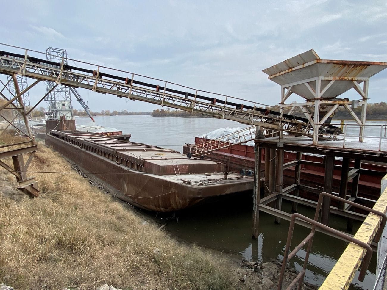 A rusted captive dock barge and a conveyor on the Missouri River bank at the AGRI-Services of Brunswick port.