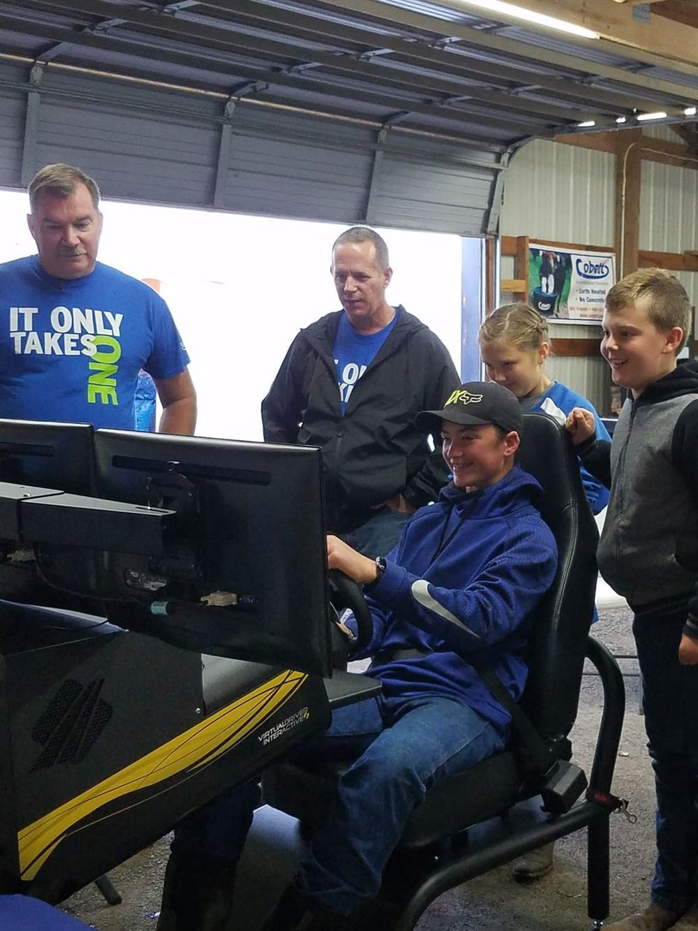 A teenager in a ball cap drives a simulator while adults in It Only Takes One shirts and two children watch.
