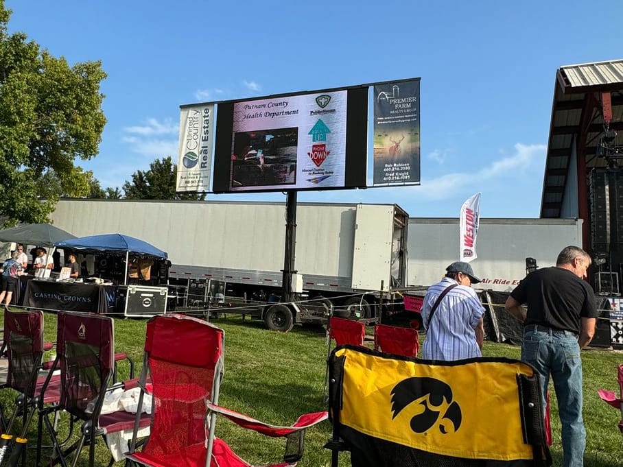 Lawn chairs face the Putnam County Fair stage, where a large screen shows a Putnam County Health Department roadway safety message.