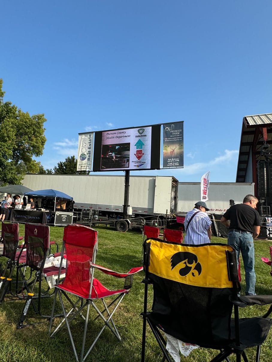 Lawn chairs face the Putnam County Fair stage, where a large screen shows a Putnam County Health Department roadway safety message.