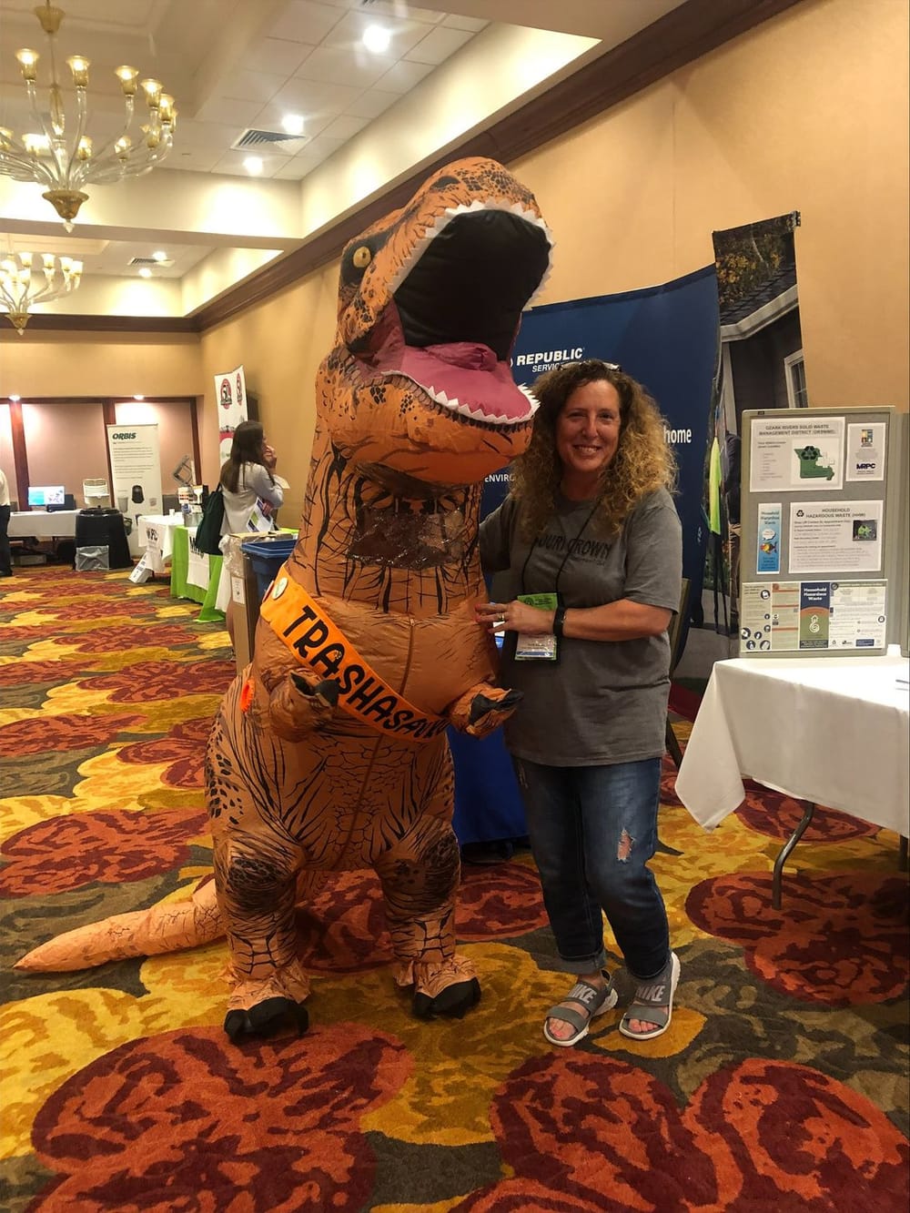 A woman poses beside a person in an inflatable dinosaur costume among exhibit tables at the Missouri Recycling Association conference.