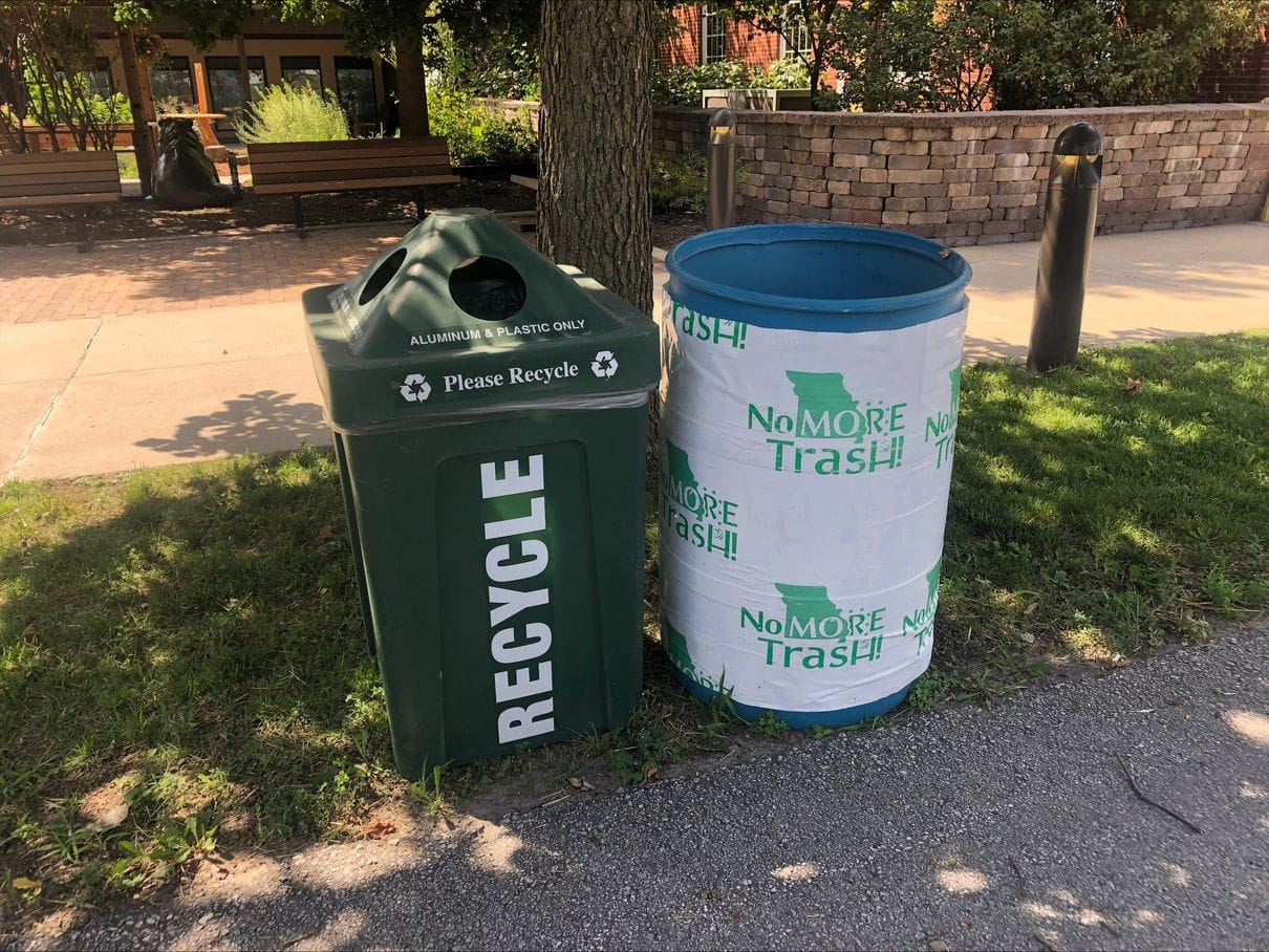A green recycling bin beside a No More Trash barrel on a shaded fairground walk.